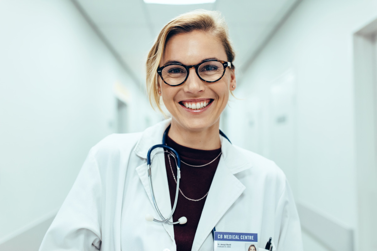 Une femme souriante, portant des lunettes et une blouse blanche ainsi qu'un stéthoscope, se tient dans un couloir blanc