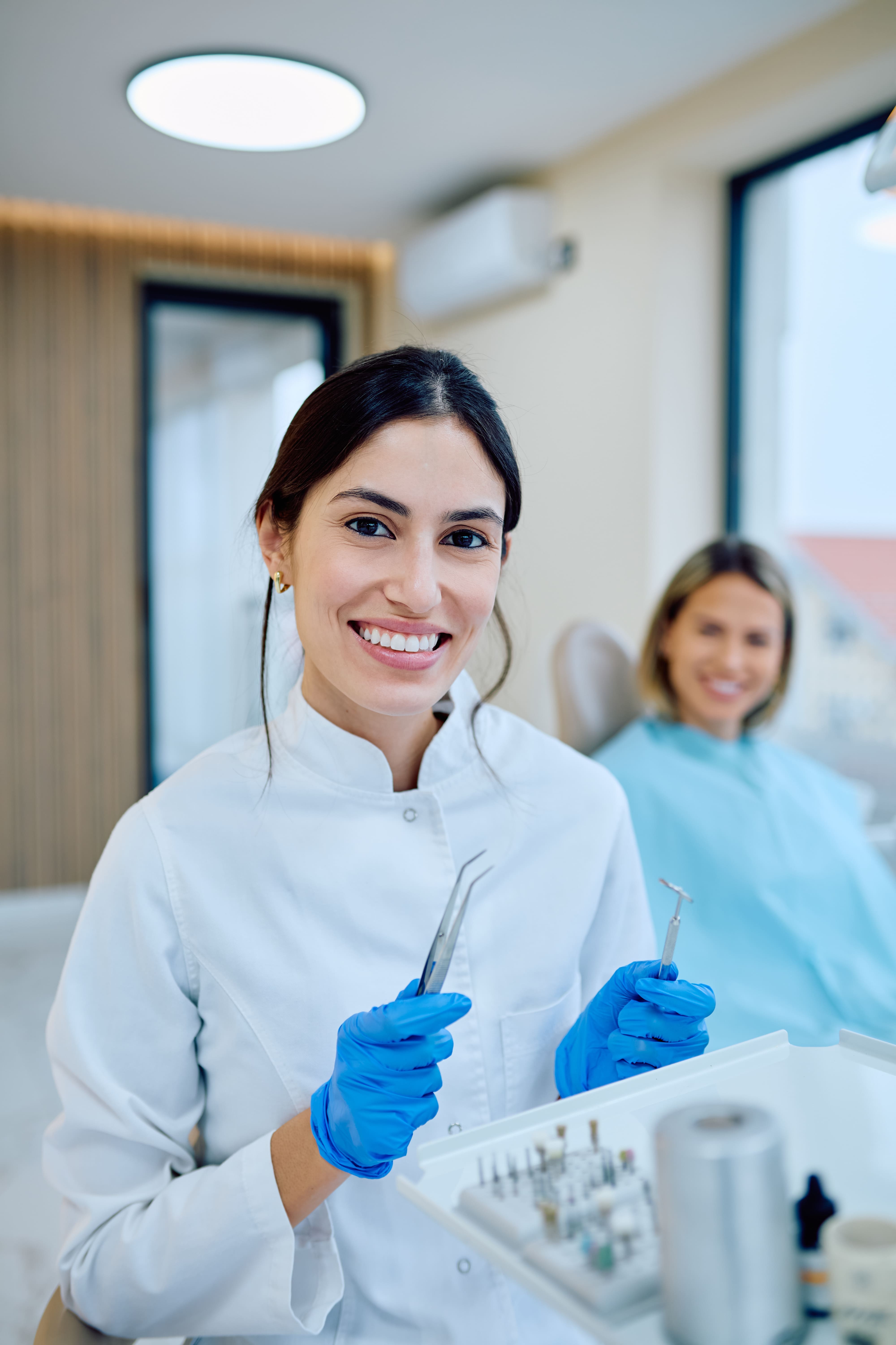 Une femme souriante, portant une blouse blanche, des gants et une pince, se tient dans un cabinet dentaire