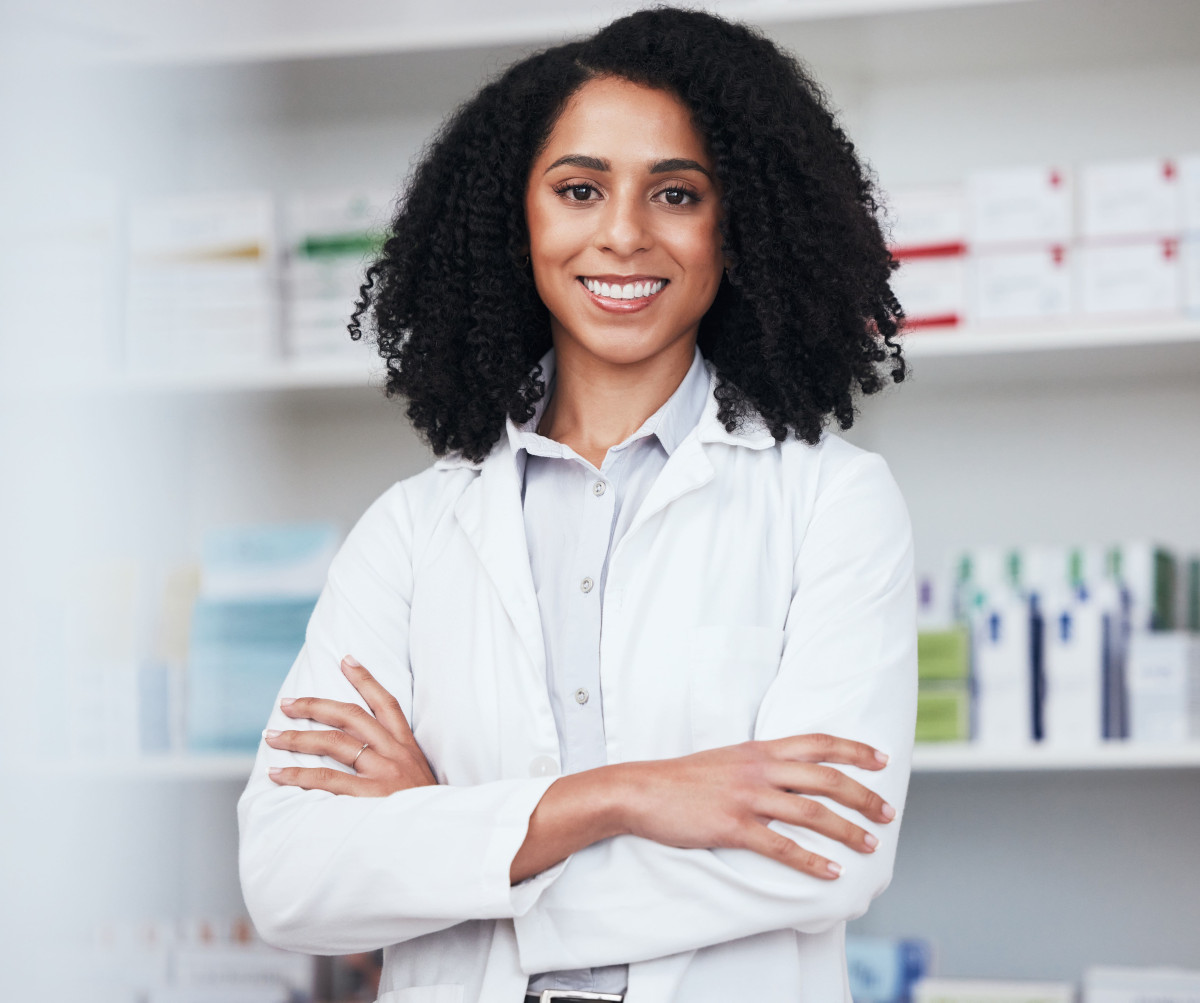 Une femme souriante, une blouse blanche, se tient dans une officine pharmaceutique